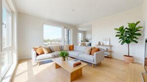 Salon spacieux avec grand canapé gris, parquet en bois clair, larges fenêtres et plantes vertes dans un appartement moderne.
